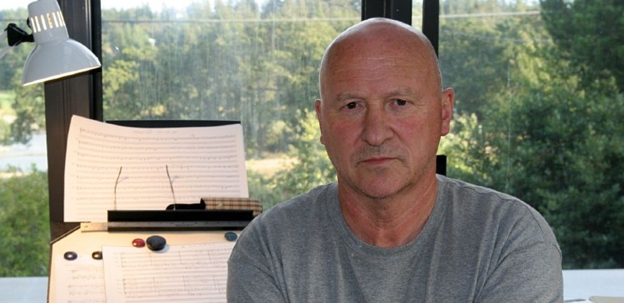 A man with a shaved head wearing a grey tee shirt sitting in front of sheet music.